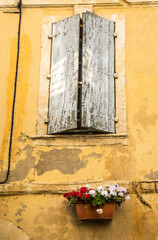 A shuttered window of a stone building with a window box of Geranium flowers in the village of Beaucaire, France.