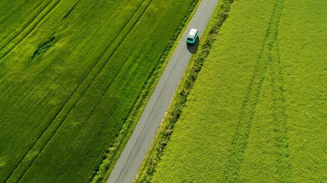 Mini bus driving on narrow country road, with green fields on both sides, drone stock footage 2