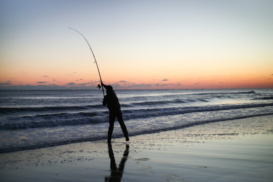 Silhouette of person surf fishing alone on ocean beach with long rod at sunset