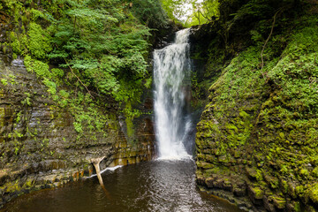 Tall waterfall in a narrow canyon surrounded by green foliage (Sgwd Einion Gam, Waterfall Country, Wales)