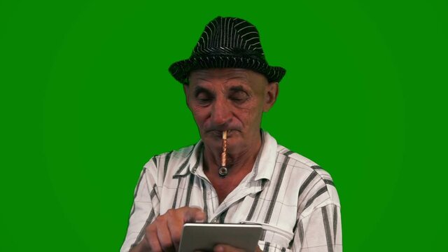 Elderly Caucasian man 70-80 years old wearing a hat smokes a pipe and browses the Internet using a tablet computer. A pensioner in an elegant hat with a digital gadget. Green screen