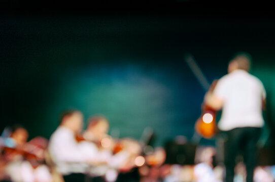 A conductor holding a violin leads a string orchestra, intentionally blurred
