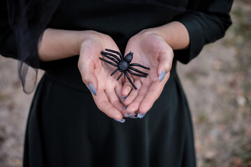 Young cute beautiful woman in dark dress and witch's hat holdsblack spider in her hands. Halloween party costume. Forest, park with autumn trees.