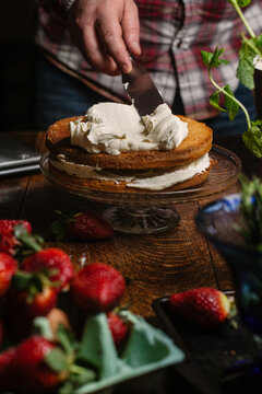 Mans hand using a palette knife to spread cream over a sponge cake.