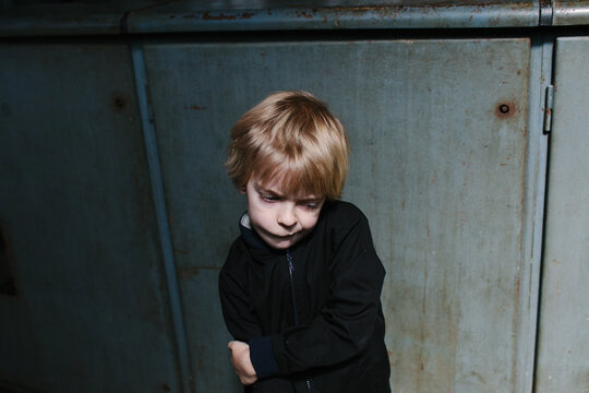 A thoughtful child looking down wearing a black jacket against grey-blue factory walls.