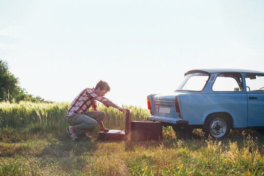 Retro-Styled Young Man Looking at Clothes in Suitcase on Vintage Car Road Trip
