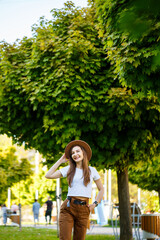 Young happy woman in a hat walks along an alley in a park. A girl of European appearance with a smile on her face on a bright sunny summer day
