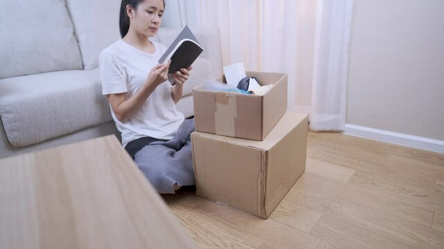 Asian pretty woman unpack parcel Storage box to arrange things to new apartment, sitting down couch behind, busy Multitasking works shore, Relocating moving in concept, stay at home, middle shot