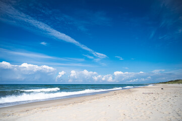 Panorama of a deserted sandy beach on which the sea foamy waves roll. An endless blue sky with white clouds on it.