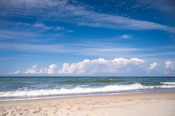 Panorama of a deserted sandy beach on which the sea foamy waves roll. An endless blue sky with white clouds on it.
