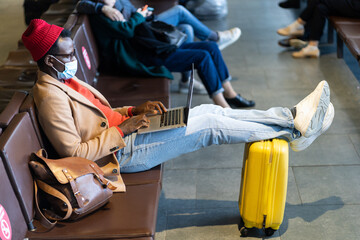 Stylish African American traveler millennial man wear face protective mask, sitting in hall of airport, works remotely on laptop while waiting for a flight and boarding. New normal. 