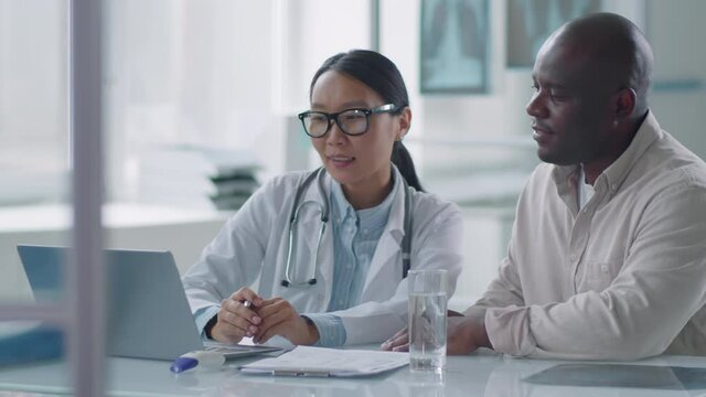 Medical worker sitting at desk with her African American colleague showing some new information about new desease on laptop to him