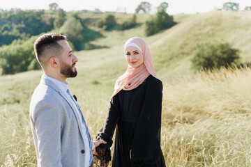 Muslim love story of mixed couple. Man and woman smiles and walks on the green hills . Woman weared in hijab looks to her man. Advert for on-line dating agency