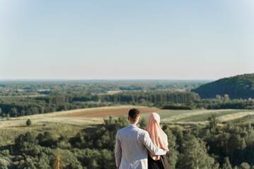 Fototapeta na wymiar Muslim love story. Mixed couple smiles and hugs on the green hills . Woman weared in hijab looks to her man. Advert for on-line dating agency