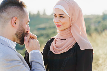 Muslim love story close-up. Mixed couple smiles and hugs on the green hills . Woman weared in hijab looks to her man. Advert for on-line dating agency