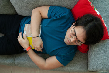 Portrait from above of a young man with glasses sleeping on a grey couch  in the living room next to the window, holding a book between his hands