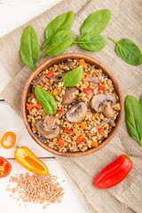 Spelt (dinkel wheat) porridge with vegetables and mushrooms in wooden bowl on a white wooden background and linen textile. Top view, close up.