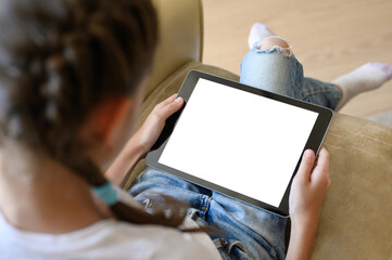 A tablet with a white display in the hands of a girl. The girl sits at home on an armchair and...