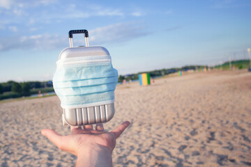 Time to travel. The concept of safe rest during a pandemic Covid-19 Coronavirus. Suitcase for travel with a medical mask on the beach in hand against the background of the beach and the sunny sky.