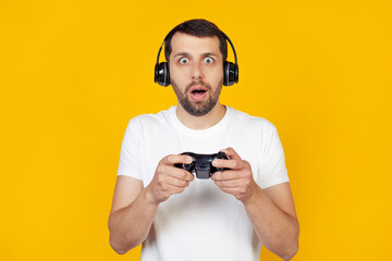 Young man with beard in white t-shirt gamer playing video games with headphones, scared in shock with surprised face, scared and agitated with fear expression Stands on isolated yellow background © Yauhen