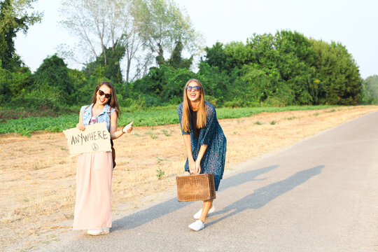 Two female friends hitchhiking with ""anywhere"" caption on the board