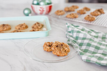 Chocolate Chip Cookies on a Plate; Cookie Sheet with Chocolate Chip Cookies on a White Countertop with Christmas Decor; Some Cooling on Wire Rack in Background