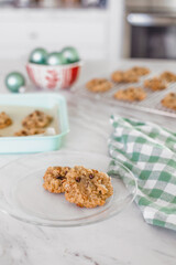 Chocolate Chip Cookies on a Plate; Cookie Sheet with Chocolate Chip Cookies on a White Countertop with Christmas Decor; Some Cooling on Wire Rack in Background
