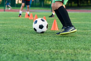 Persistent teen boy in sportswear trains football on soccer field and learns to circle the ball between training cones.