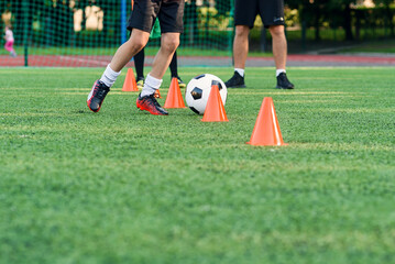 Obraz premium Close up boy in sportswear trains football on soccer field and learns to circle the ball between training orange cones.
