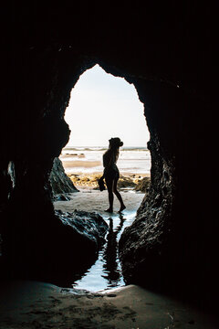 A Young Woman Stands In Wonder Outside The Mouth Of An Ocean Carved Cavern On The Beach