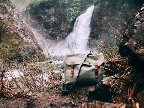 Old Swiss Army Bag With Leather Buckles On Ground In Front Of Flowing Waterfall
