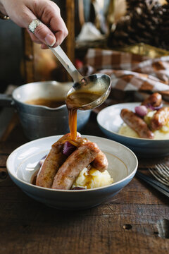 Woman's hand pouring gravy over a bowl of bangers and mash.