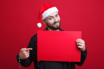 Young man with beard and new year hat is pointing to red blank space on a banner holding on a studio wall