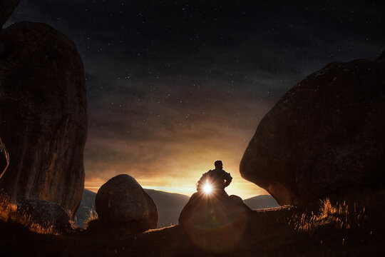 A young astronaut sits and ponders under the setting sun as a starry night emerges above him.