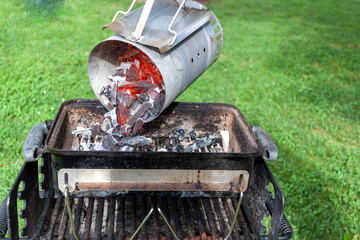 close up image of charcoal chimney starter filled with red glowing charred coal pieces. The chimney is lifted up from its handle and red hut burning coal pieces are poured into the grill