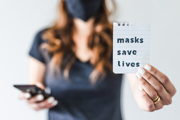 covid-19 pandemic outbreak, woman holding Masks save lives text towards the camera while browsing on her smartphone