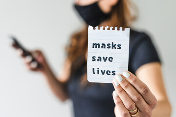 covid-19 pandemic outbreak, woman holding Masks save lives text towards the camera while browsing on her smartphone