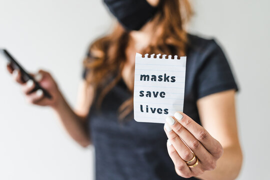 covid-19 pandemic outbreak, woman holding Masks save lives text towards the camera while browsing on her smartphone - Powered by Adobe