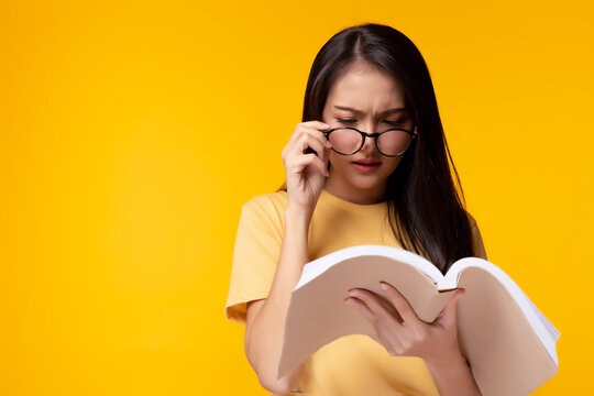 Young woman can not read book because her eyesight's getting worse. Young Asian girl wears eyeglasses and holding text book. She is university student. Isolated on yellow background, copy space