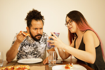 Pareja de novios comiendo pizza en casa viendo telefono inteligente