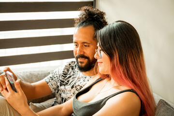 Joven pareja mexicana pasan tiempo juntos en sillon dentro de casa utilizando un telefono inteligente