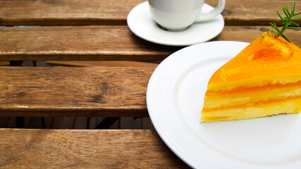 Close up an orange cake topping with a sliced piece of fresh orange in white dish or plate on wooden table with copy space on left and blurred white cup of coffee. Relax with dessert and hot drinking
