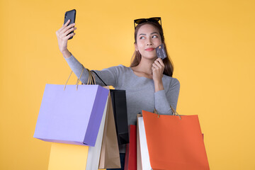 Beautiful young Asian woman with colourful shopping bags with credit card and taking selfie with smartphone isolated on yellow background