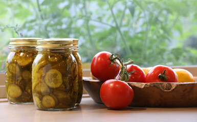 Three full freshly preserved Dill Cucumber gherkin jars alonside plump ripe vine tomatoes in a beautiful wooden fruit bowl. Home made preserving pickling cooking in the kitchen concept