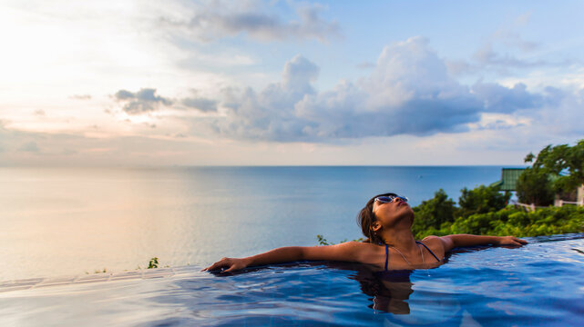 Woman relaxing at the edge of swimming pool with sea horizon and sky in background during sunset.