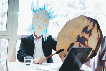 Asian businessman with tambourine in shamanic mask. Concept of attracting success, wishing good luck to business. Money increasing. Occult ritual, spiritism. Sitting at the table with laptop.