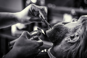 Barber works with a beard clipper. Hipster client getting haircut. Hands of a hairdresser with a beard clipper, closeup. Black and white - obrazy, fototapety, plakaty
