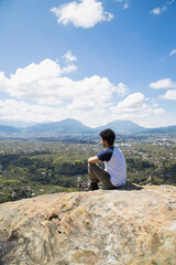 Fototapeta premium Young hispanic man sitting on top of the mountain watching the horizon - man meditating in the heights observing the volcano and mountains in front of him