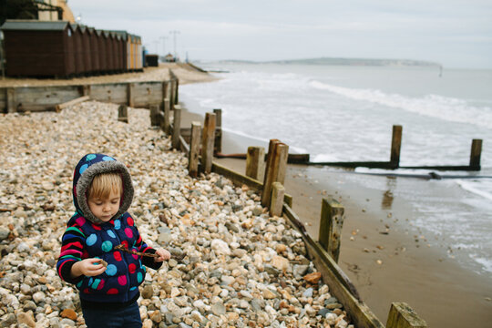 Little girl wearing colourful dots and stripes plays on a pebble beach in winter.