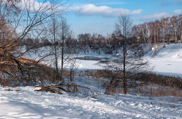 Early spring, all in the snow, thawed patches on the river. The path has high red grass. On the opposite bank, there are village houses. Blue sky with clouds.